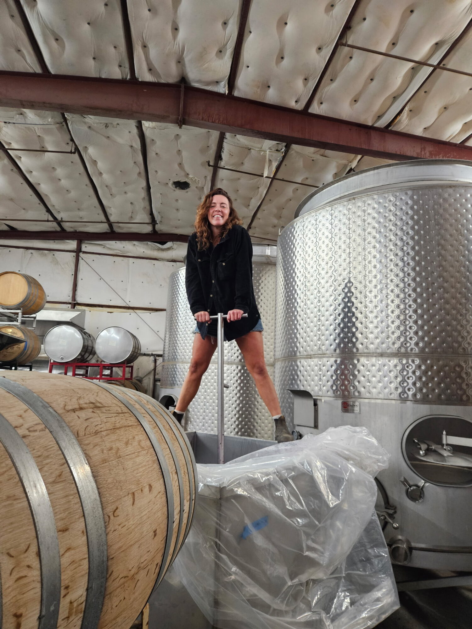 Person standing on a barrel in a winery or brewery, surrounded by large metal tanks and barrels, under a curved ceiling.