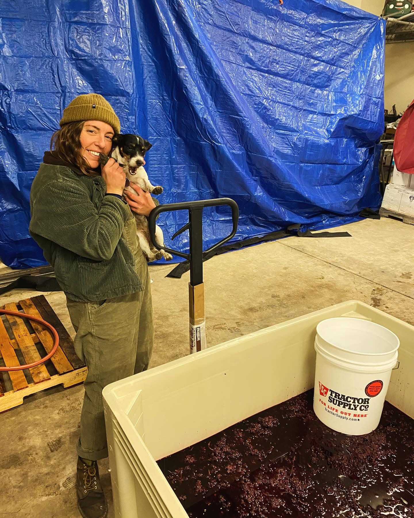 Woman in green jacket and hat holding a small black and white dog, standing in front of a blue tarp in a garage.