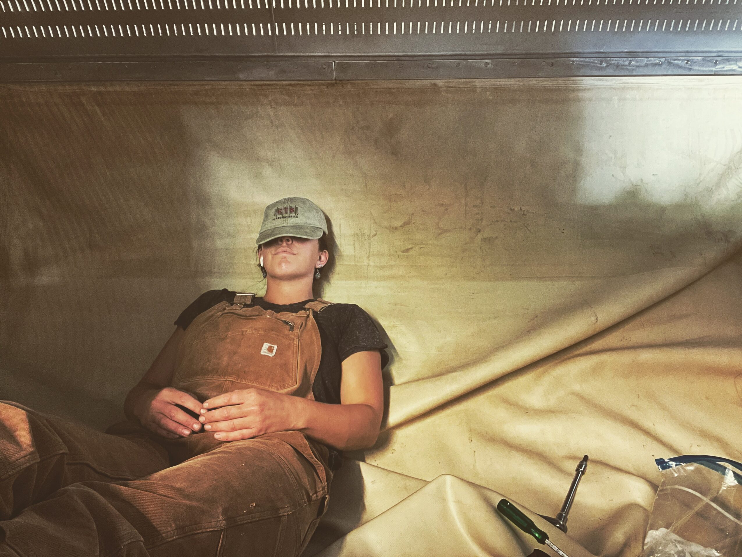 Person lying down wearing a cap, black shirt, and brown apron, with hands clasped, against a beige background.