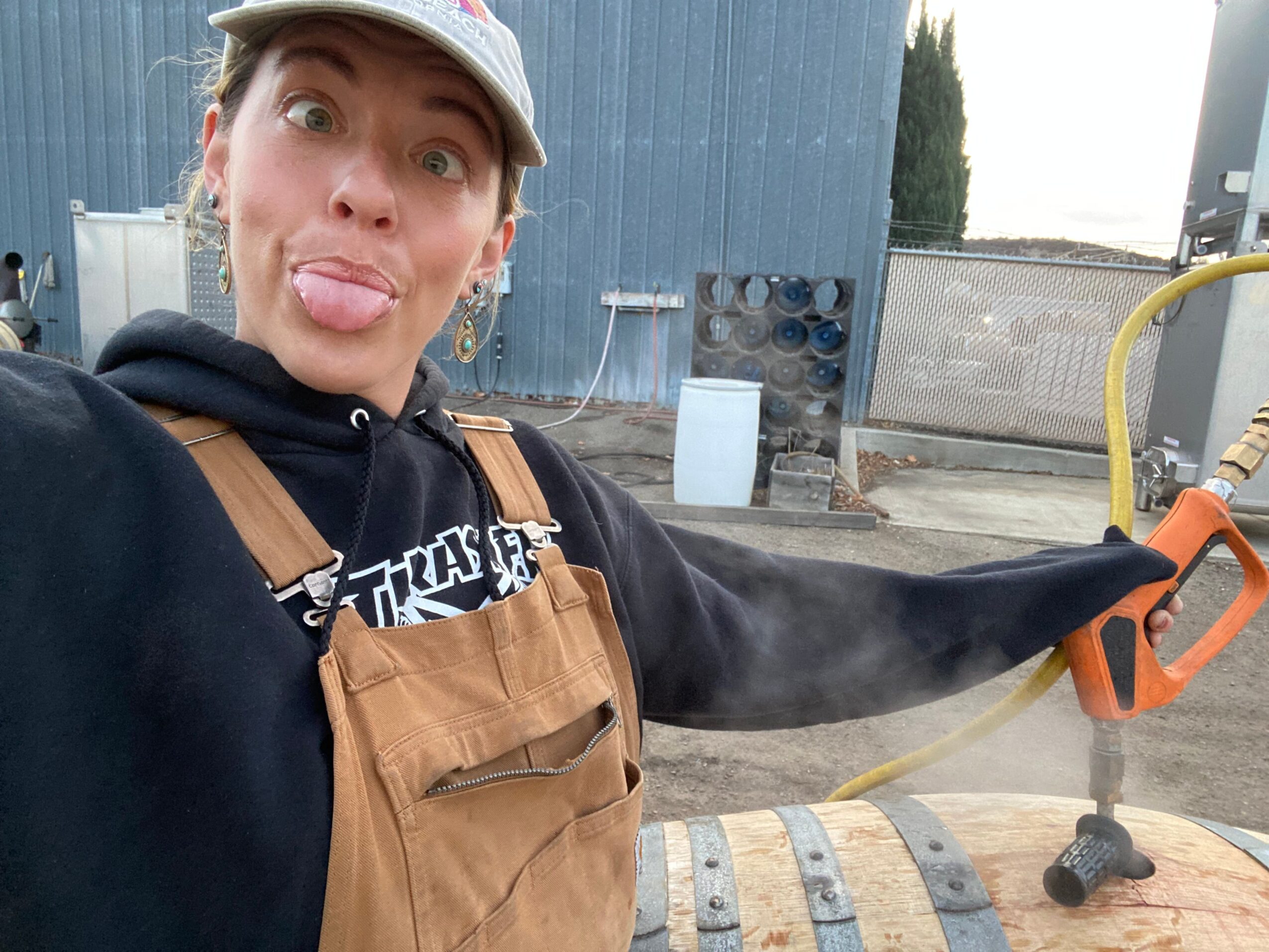 Person sticking out tongue, wearing a cap and overalls, holding a power tool outdoors near a blue building.