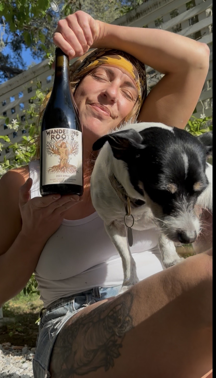 Person with yellow headband holding a dog outdoors, holding a bottle labeled 'Wander Root,' with trees and a building in background.