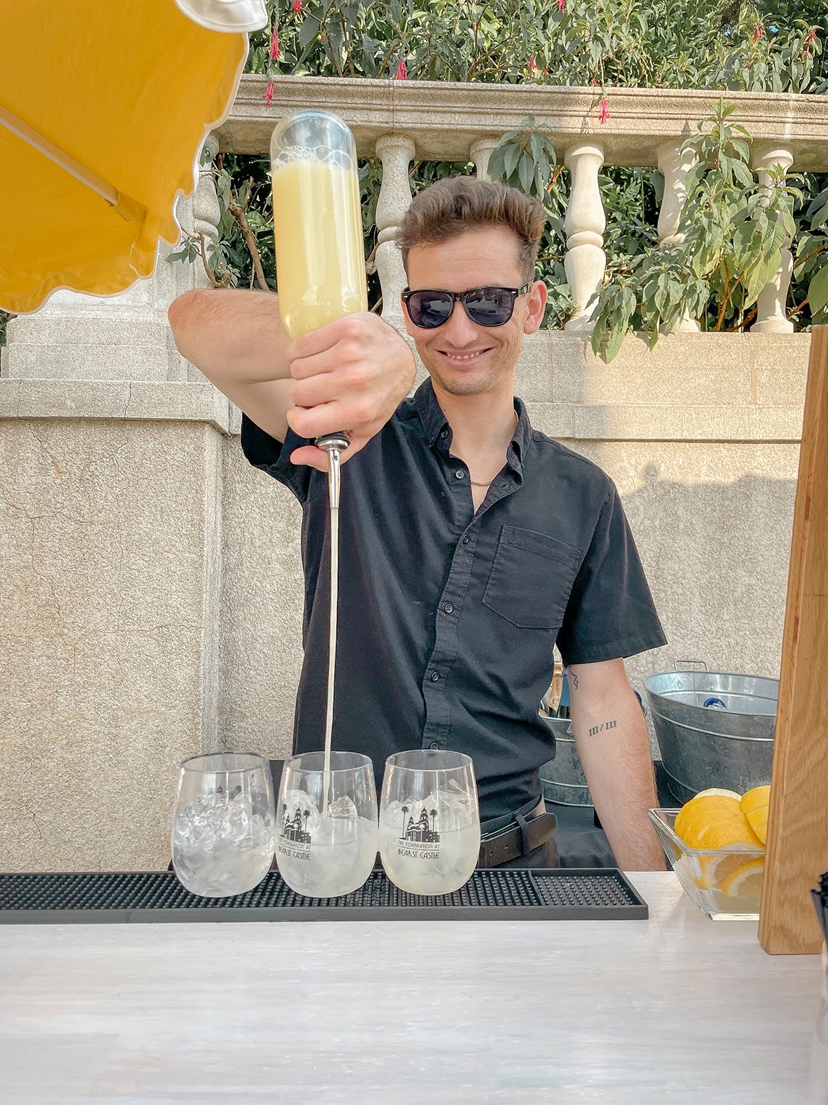 Young man wearing sunglasses and a black shirt pours a yellow drink into glasses outdoors.