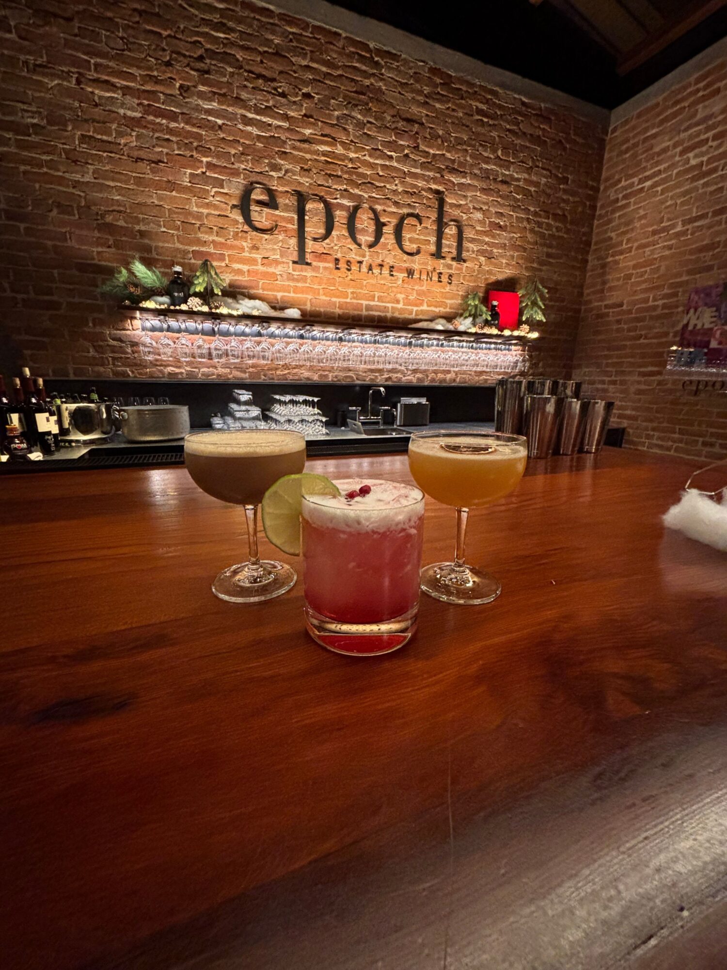 Three colorful cocktails on a bar counter with a brick wall and a sign reading 'epoch' in the background.