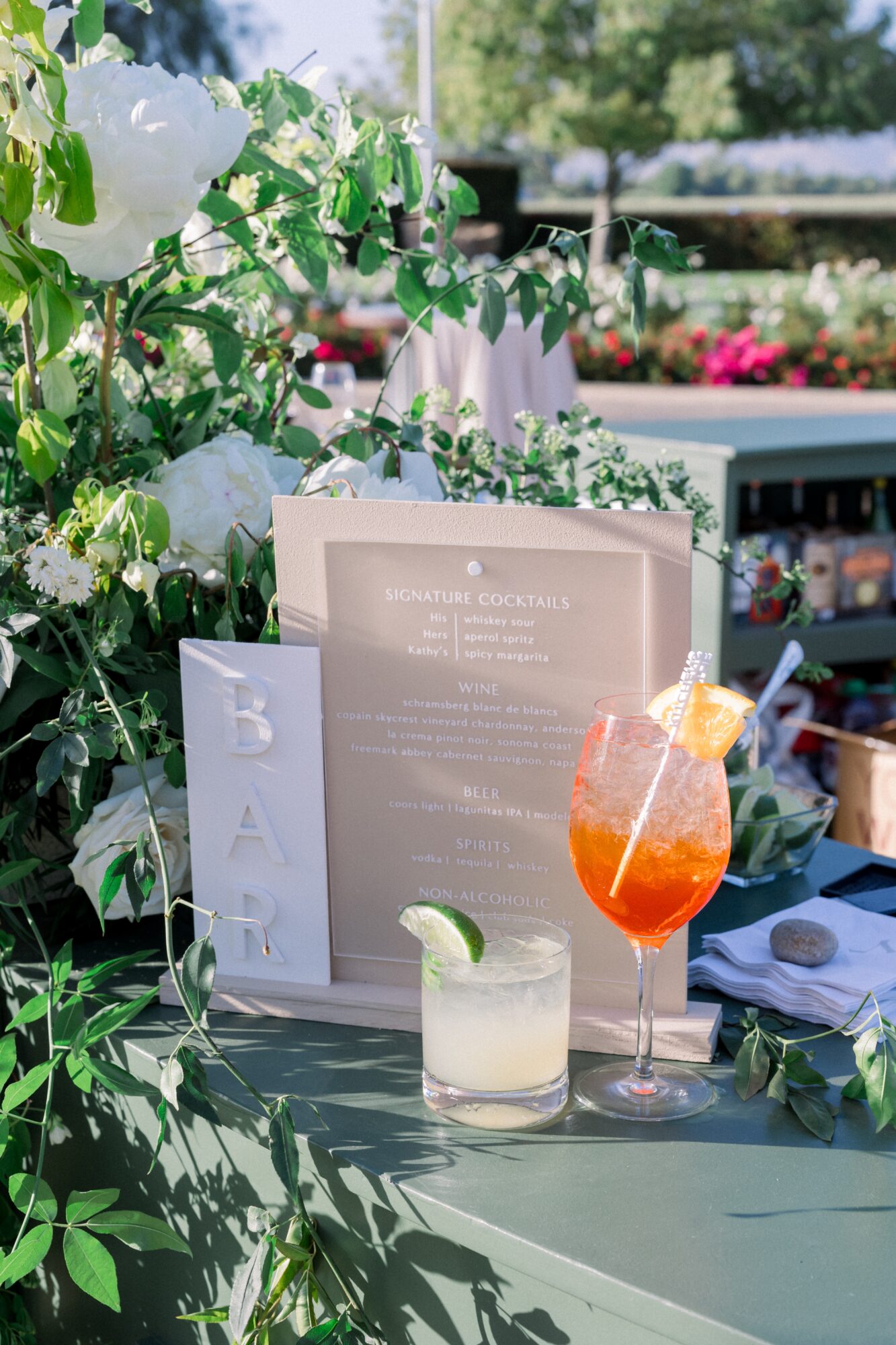 Outdoor bar with drinks, menu, and white flowers, sunny day with trees and flowers in background.