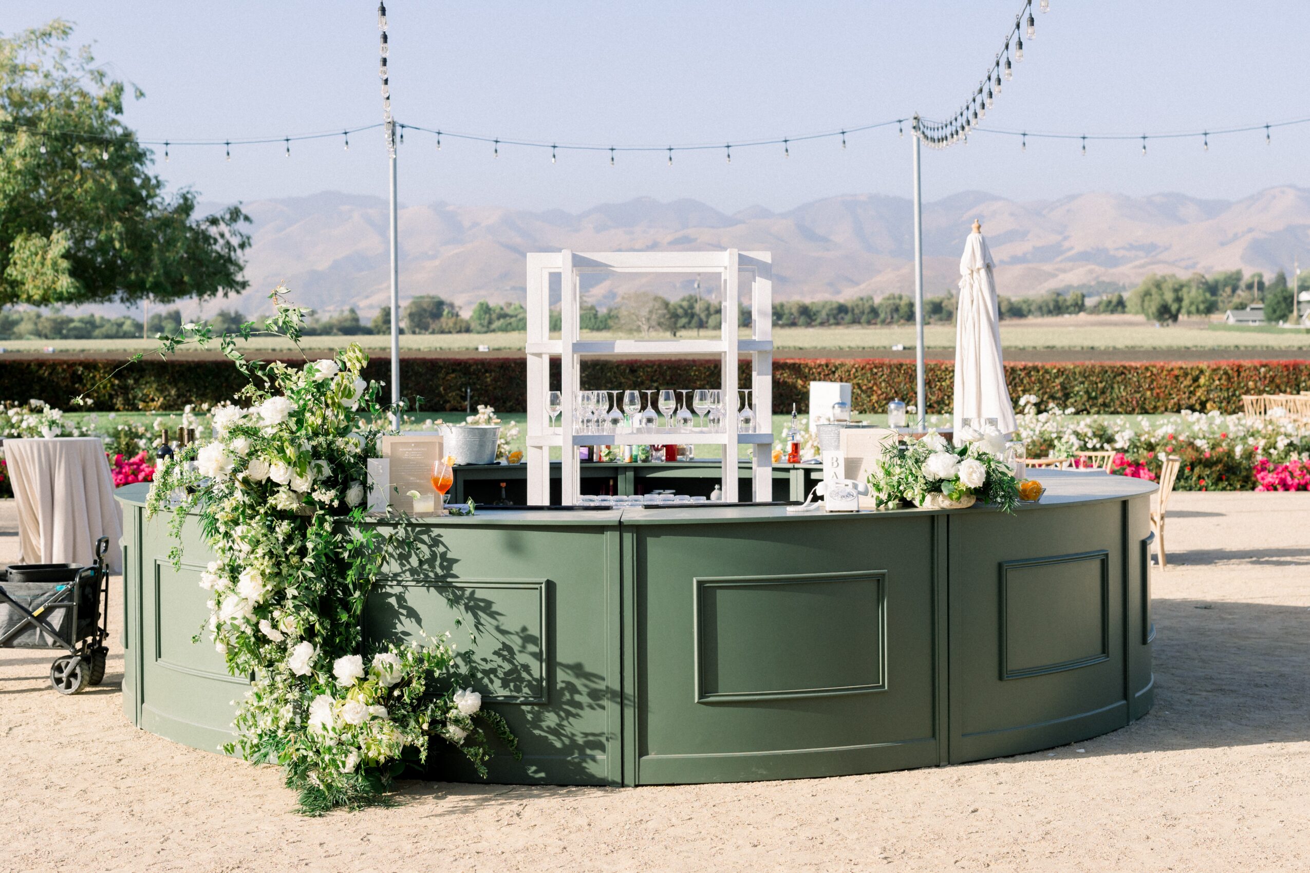 Outdoor bar with white flowers and drinks, set against a landscape with mountains and string lights, under a clear sky.