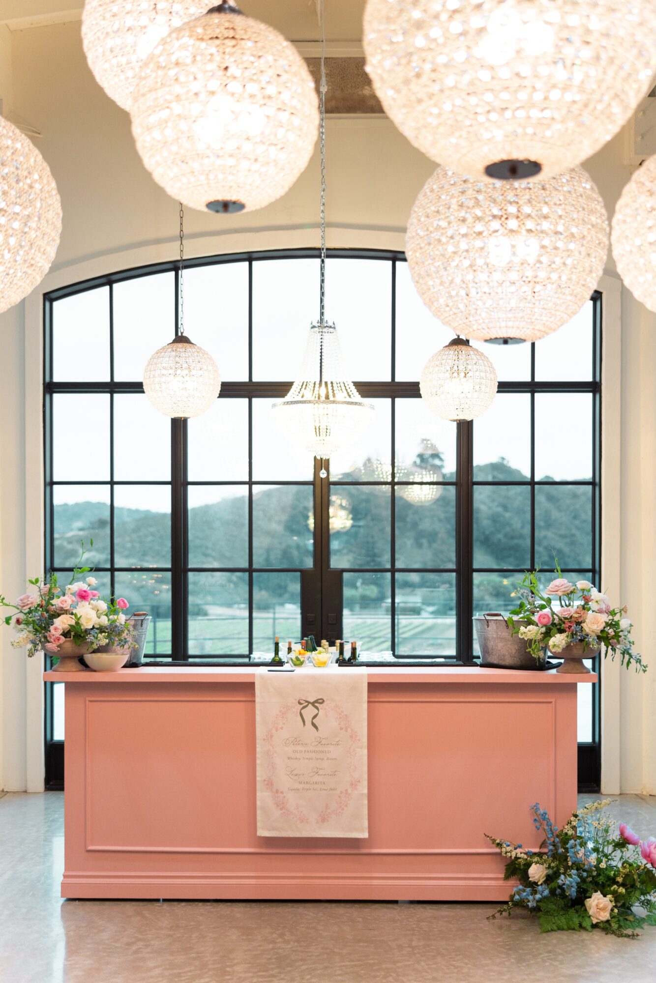 Pink counter with flowers, large window with view of trees, hanging white spherical lights, and a beige cloth on the counter.