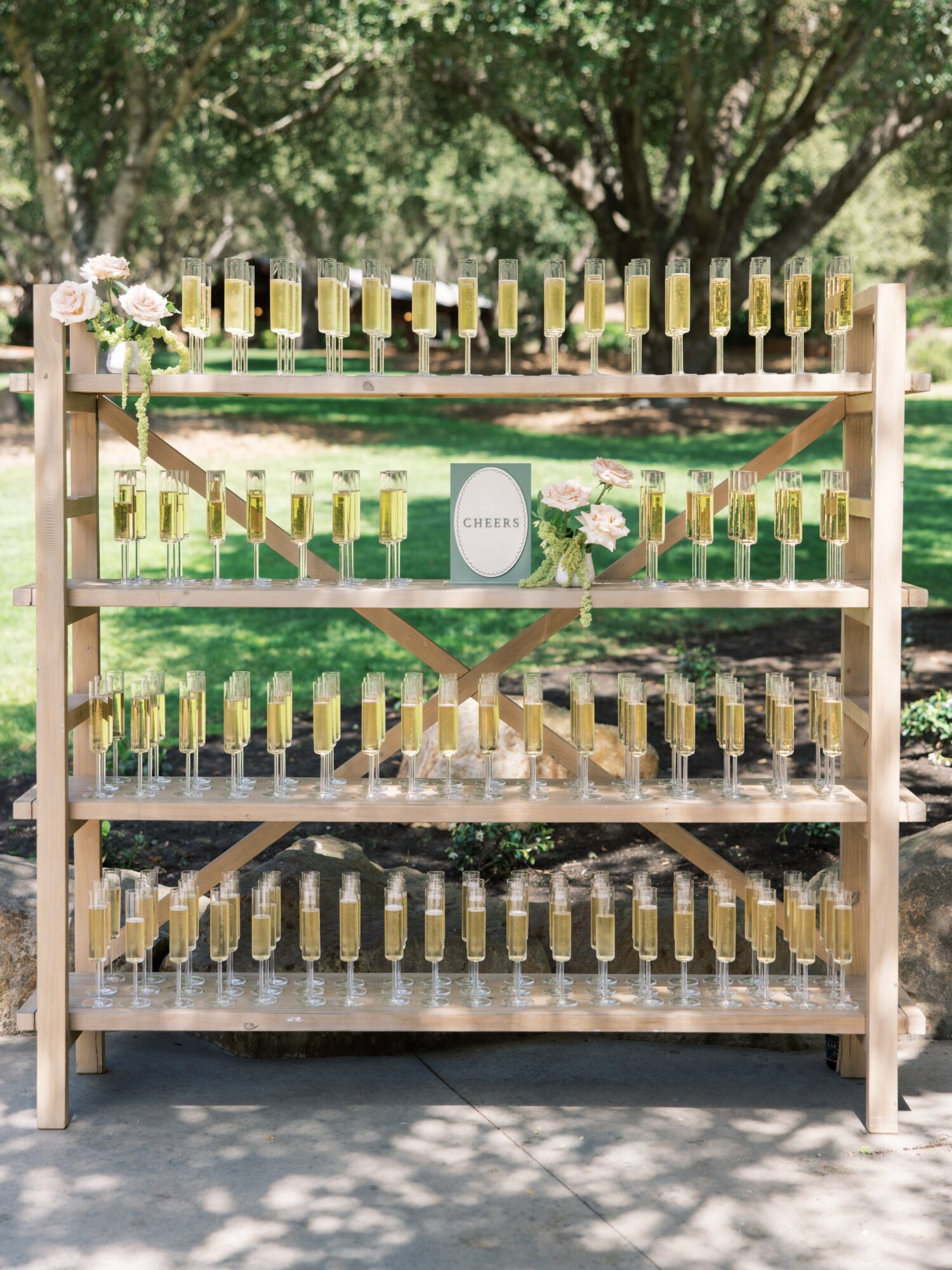 Wooden display shelf with multiple rows of yellow bottles, decorated with flowers, outdoors with trees and grass in background.