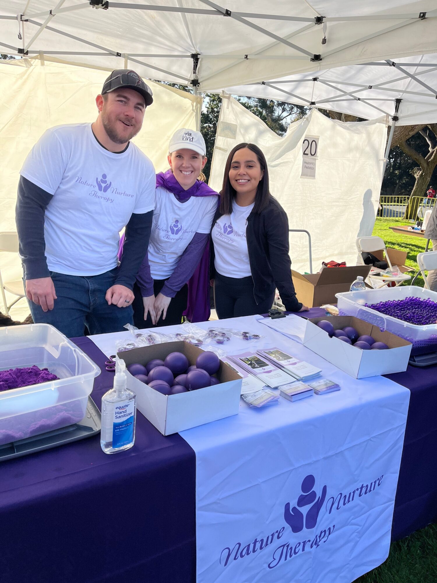 Three people standing behind a table with purple eggs and informational materials under a white canopy, outdoors.