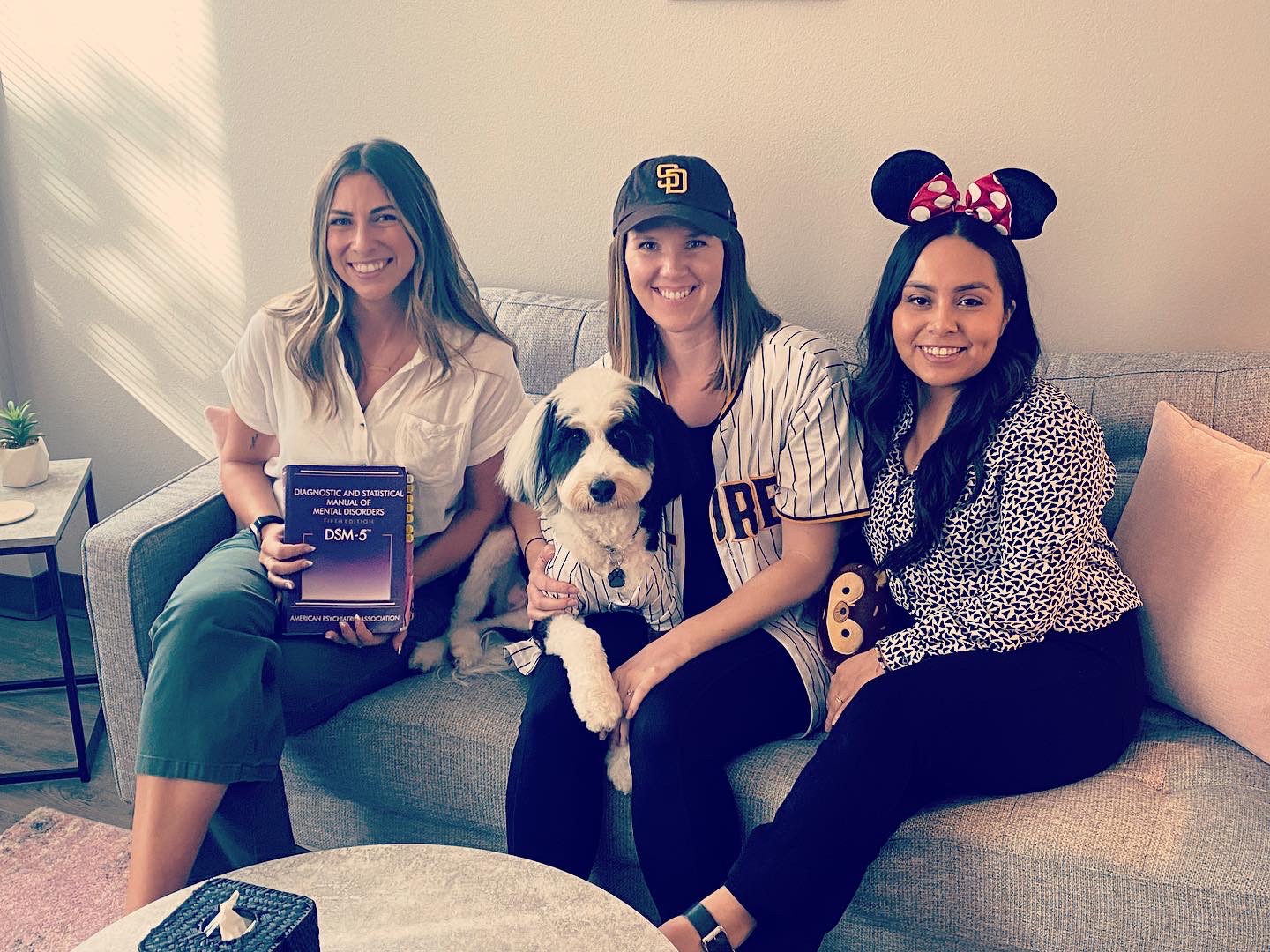 Three women and a dog sitting on a sofa in a living room, smiling at the camera.
