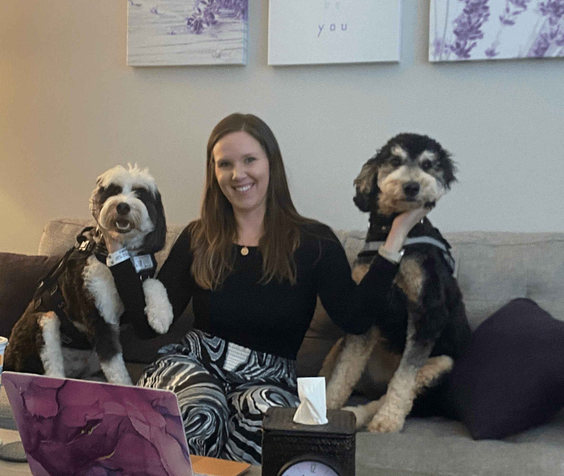 Woman sitting on a couch with two dogs, one on each side, in a living room. Wall art is visible behind.