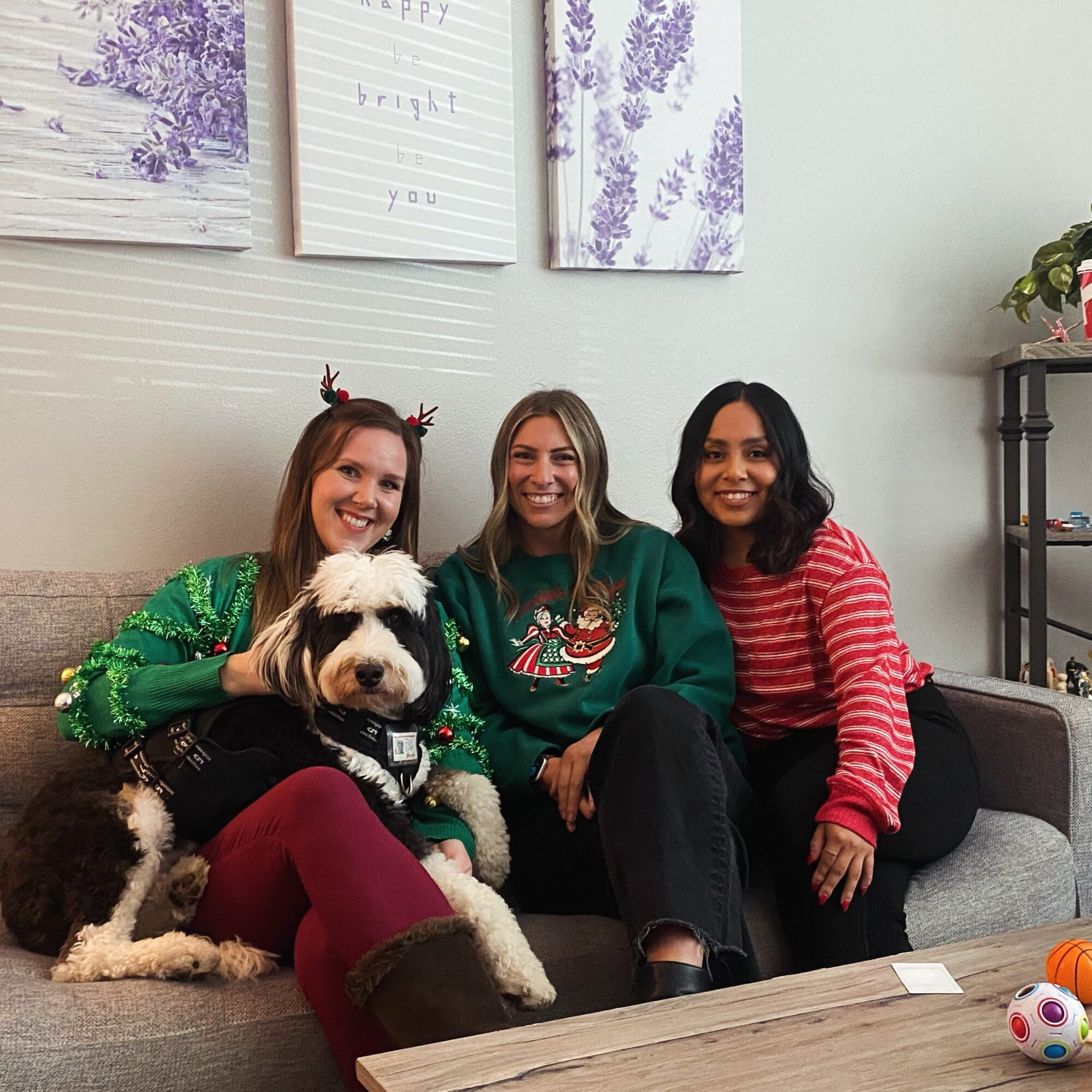 Three women sitting on a couch with a dog, smiling, in a decorated room with lavender artwork on the wall.