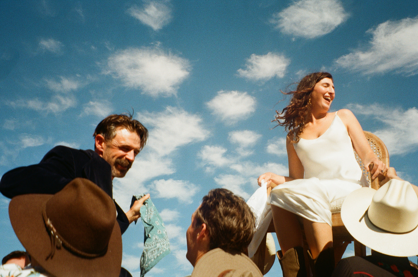 Group of people outdoors under a blue sky with clouds, smiling and talking, some wearing hats.