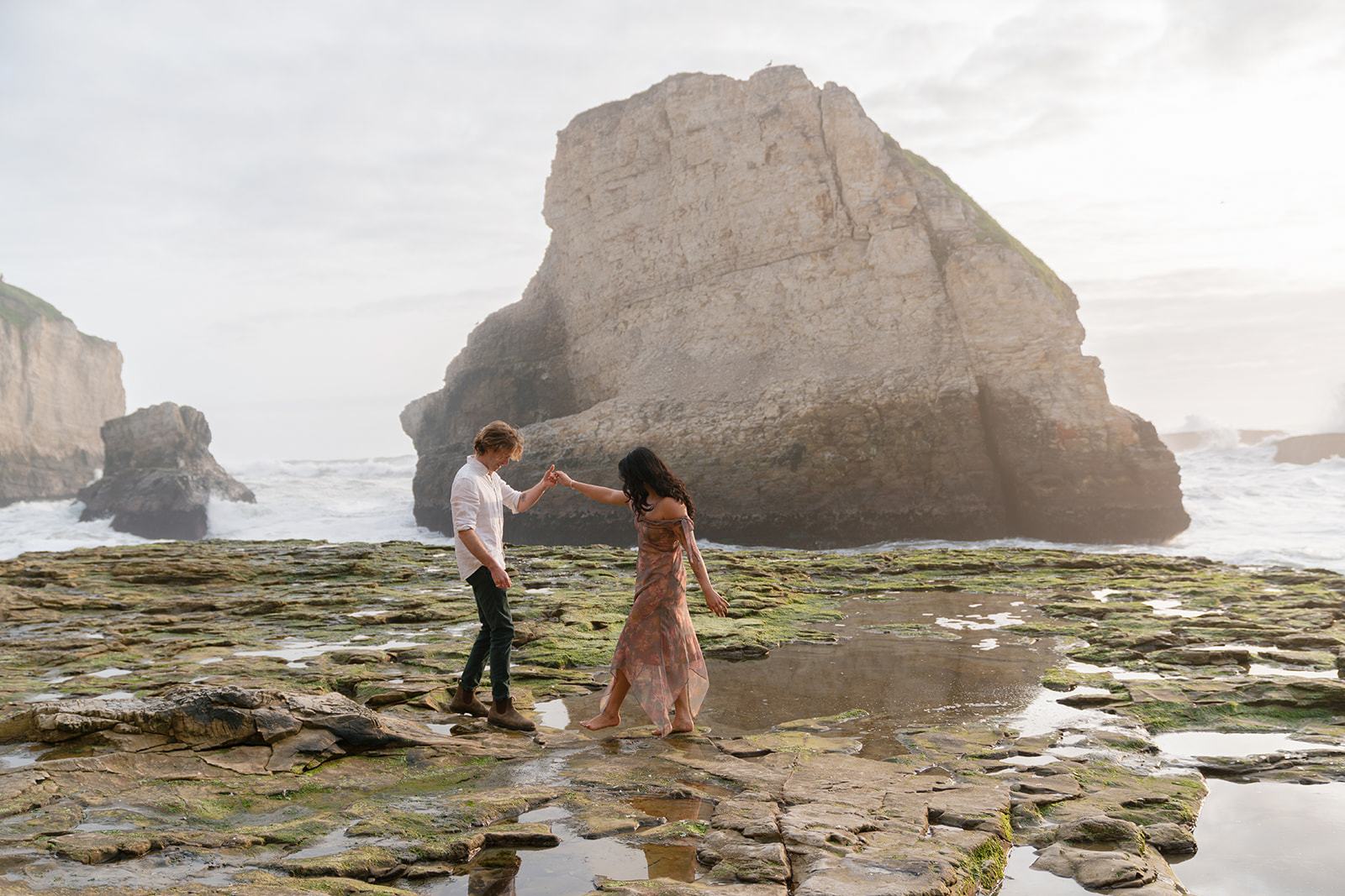 Two people stand on rocks near the water, holding hands with large rock formations in the background.