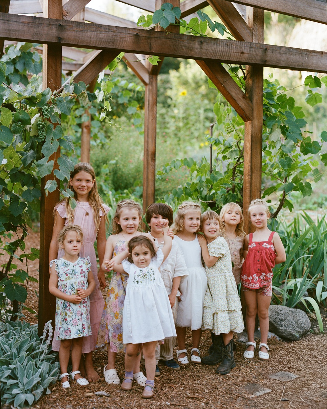 Group of nine young girls standing outdoors under a wooden structure with greenery around them.