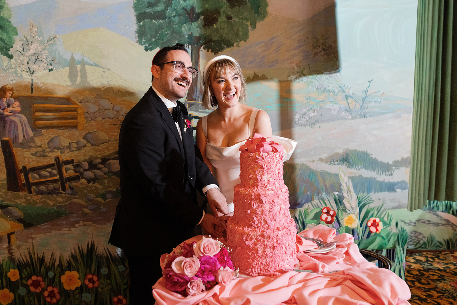 A newlywed couple in wedding attire stands beside a pink wedding cake with flowers, in front of a painted outdoor scene backdrop.