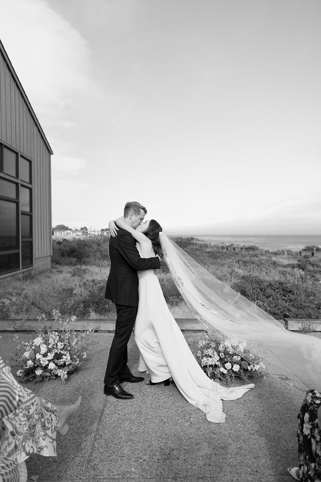 Couple in wedding attire embracing outdoors near a building, with a landscape and sky in the background.