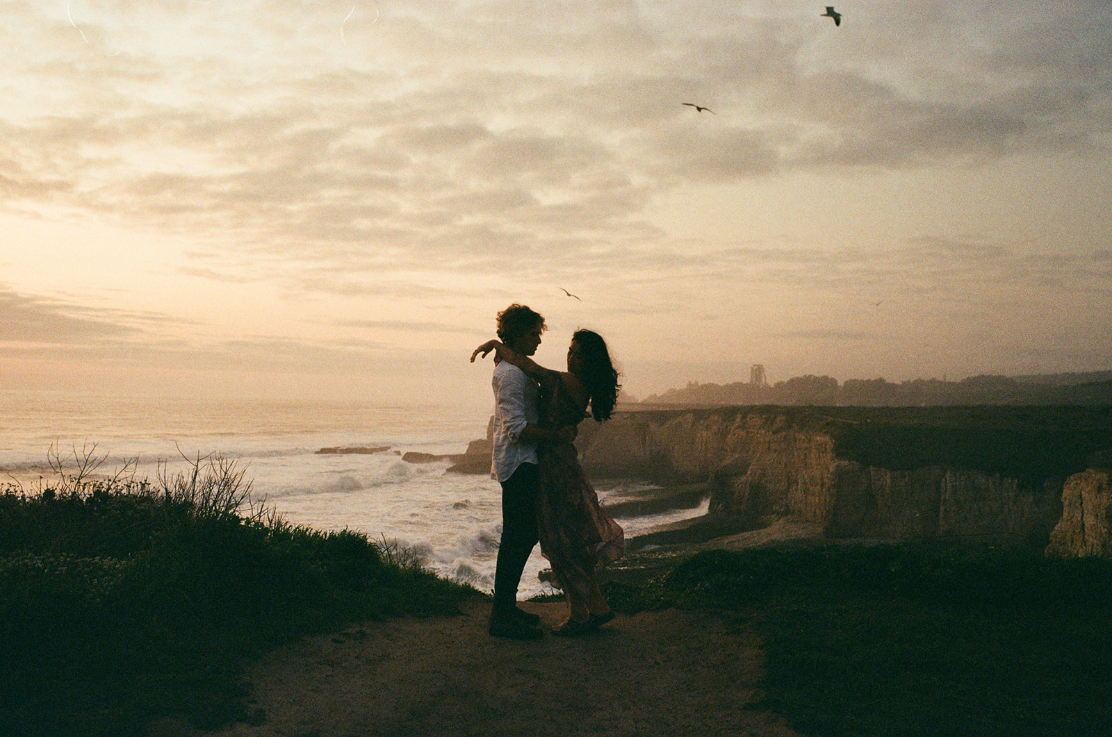 Two people stand close together on a coastal cliff at sunset, with the ocean and sky in the background.