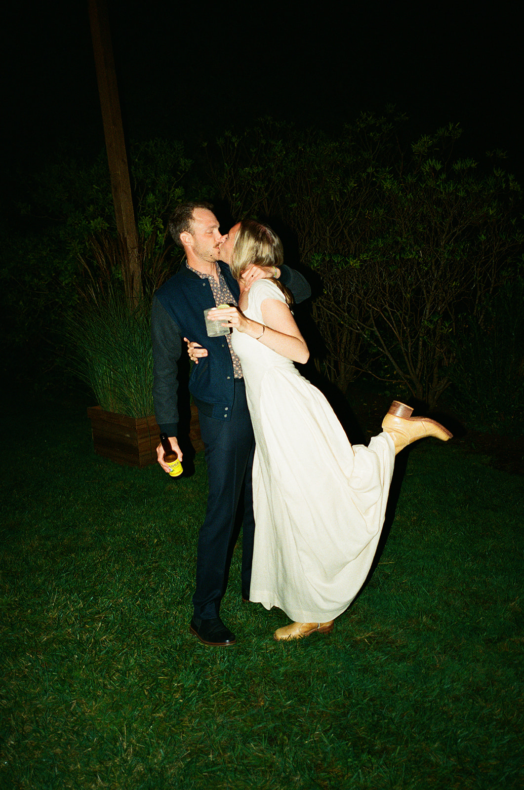 Two people, one in a dark suit and the other in a white dress, share a kiss outdoors at night.