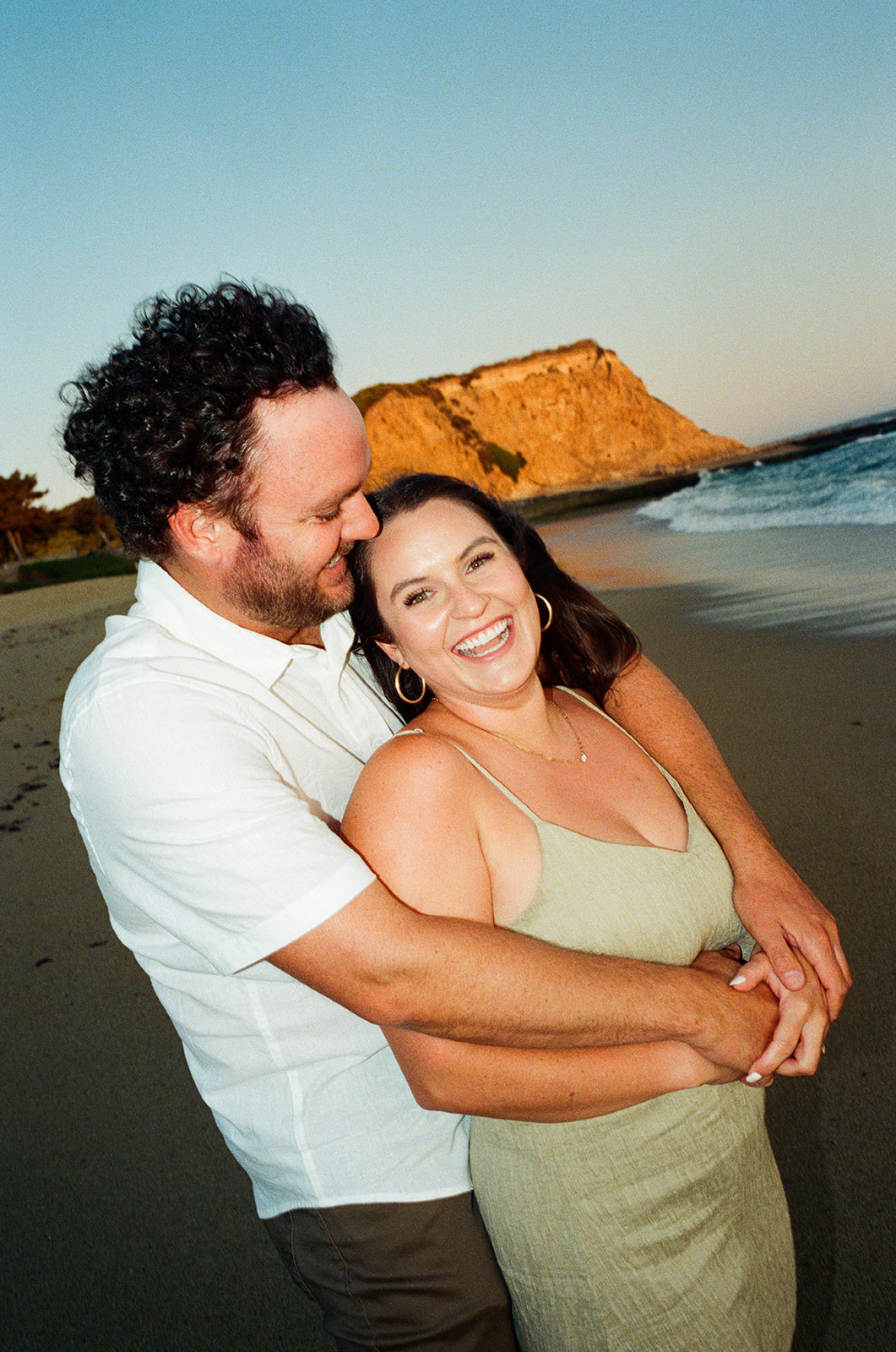 A smiling couple hugging on a beach with cliffs and ocean in the background during sunset.