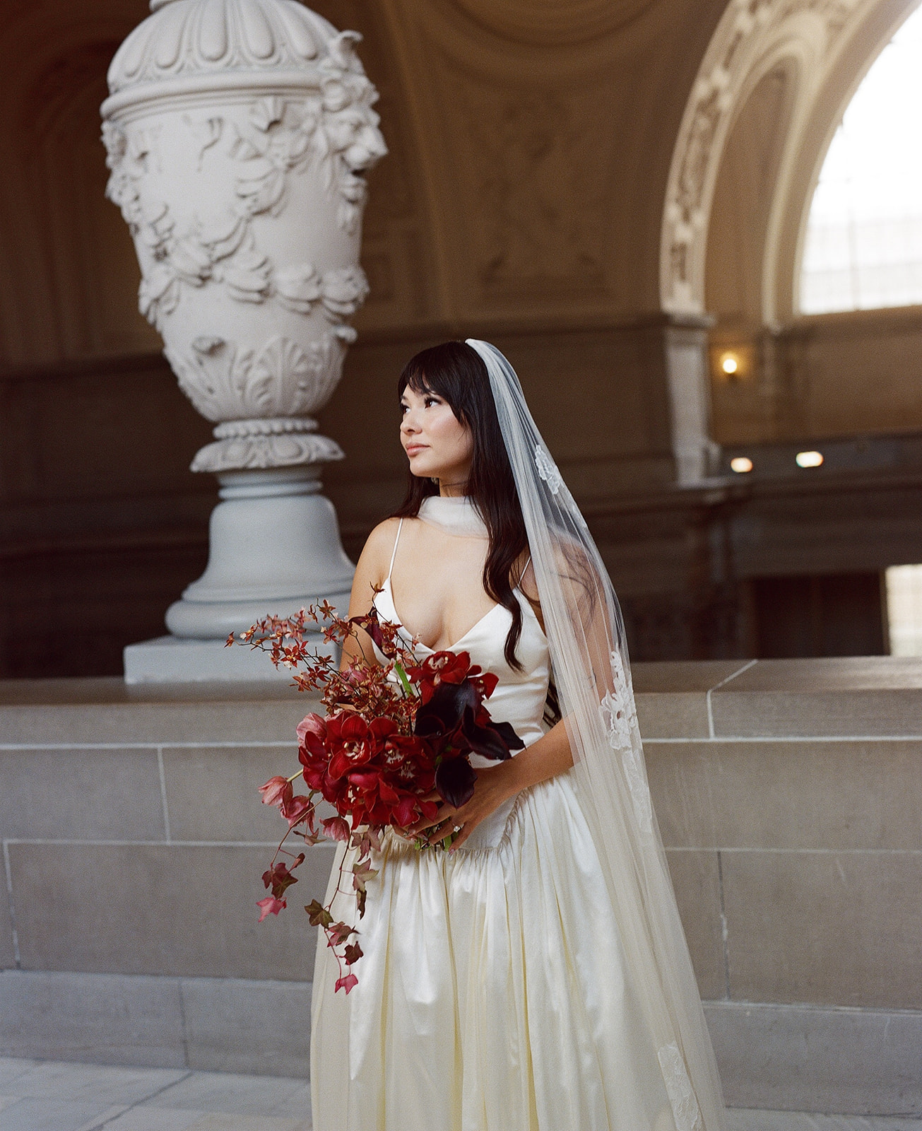 Bride in wedding dress holding a bouquet of red flowers, standing indoors near a large decorative urn and arched window.