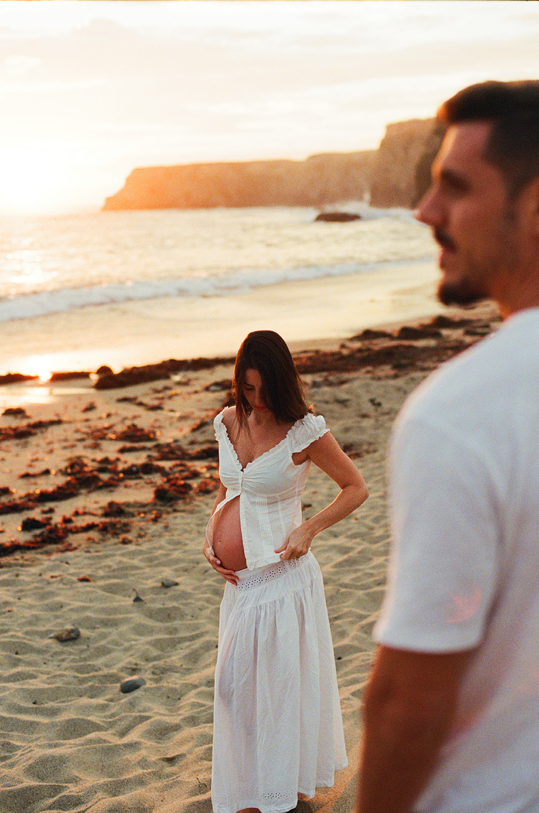 Pregnant woman in a white dress standing on a beach, with a man partially visible in the foreground, during sunset.