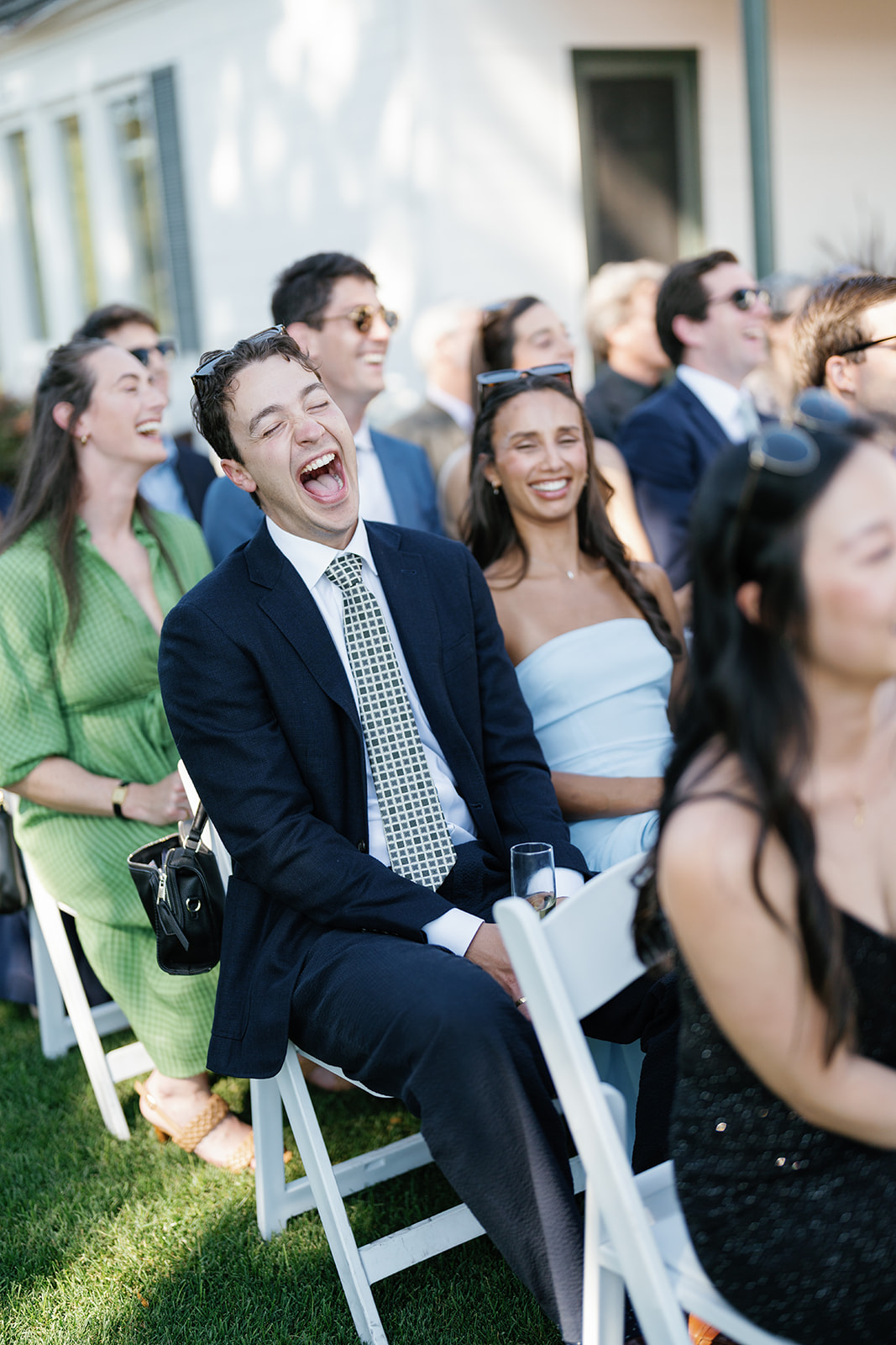 Group of people outdoors, smiling and laughing, seated on white chairs, dressed in formal attire, sunny day.