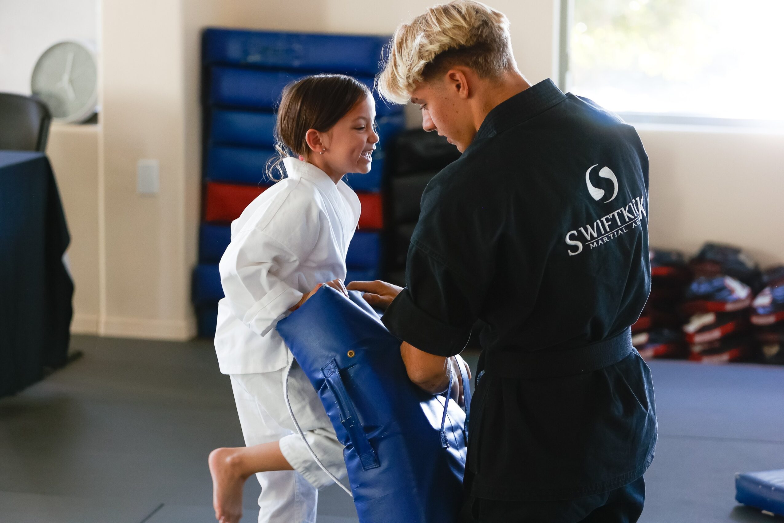 A young girl in a martial arts uniform receives a blue belt from an instructor in a training room.