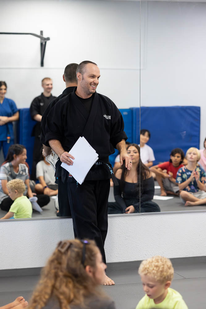 Man in martial arts uniform holding papers, smiling, in a gym with children and adults watching, mirror in foreground.
