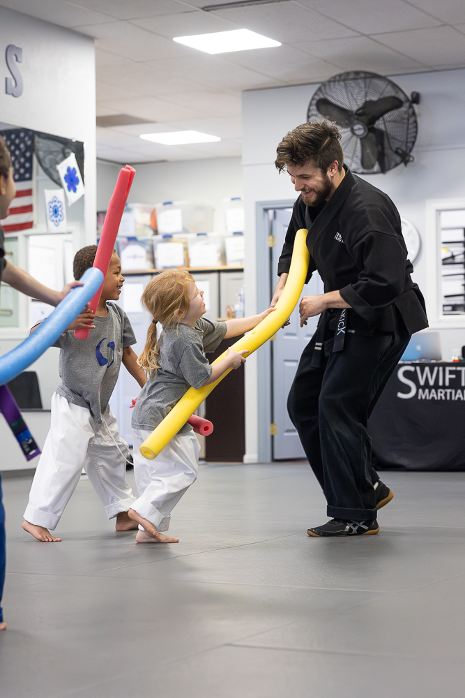 Children and an instructor holding foam noodles in a martial arts class, practicing with a large foam sword, in a training room.