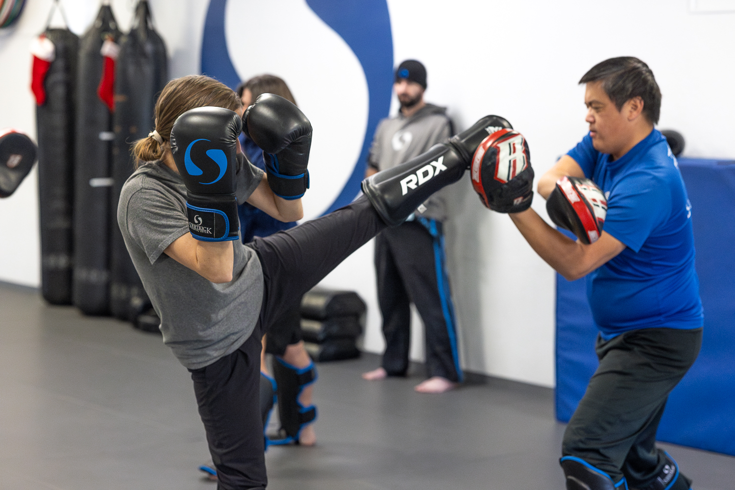 Two people practicing boxing in a gym, one person throwing a kick while the other blocks with gloves.