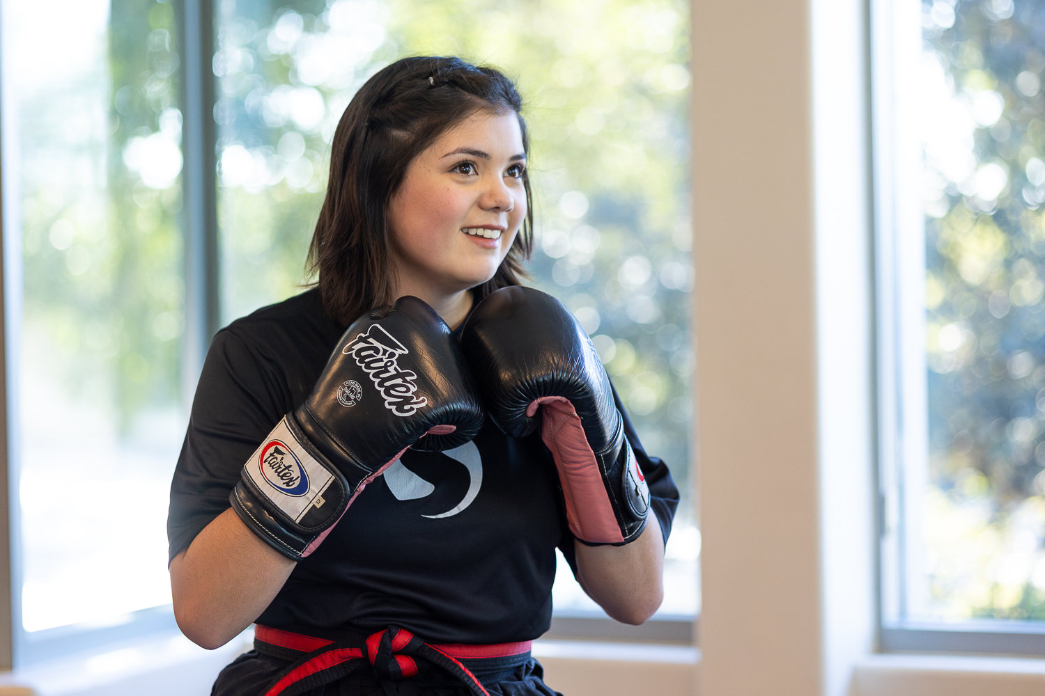 Young woman in boxing gloves smiling indoors near large windows, with trees outside visible.