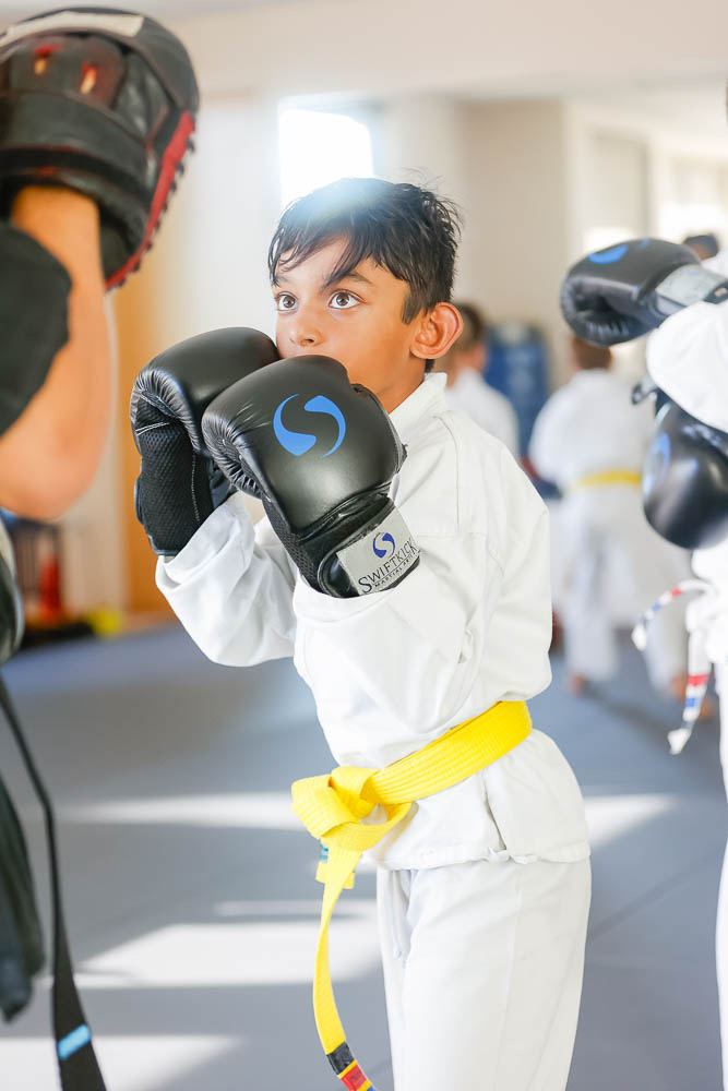Young boy in a martial arts uniform with a yellow belt practicing boxing with an instructor, wearing black gloves.
