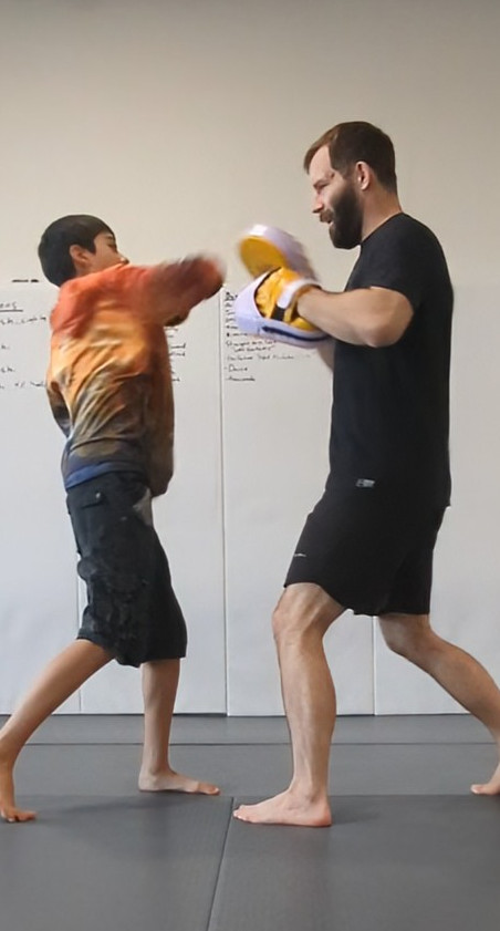 Two people practicing boxing, one young boy and an adult man, in a gym with white walls and a black floor.