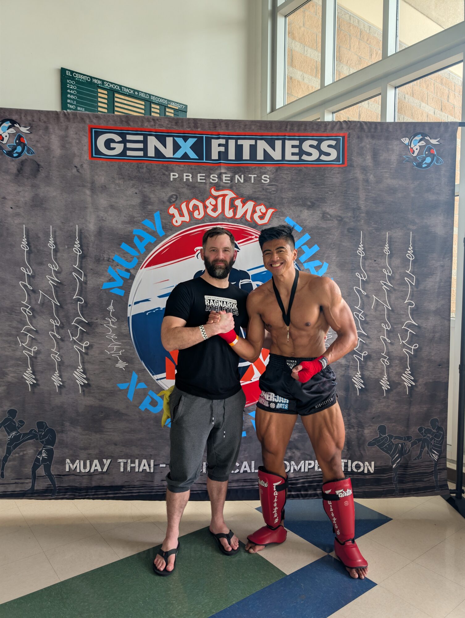 Two men pose in front of a fitness event backdrop, one in casual clothes and the other in Muay Thai gear, smiling.