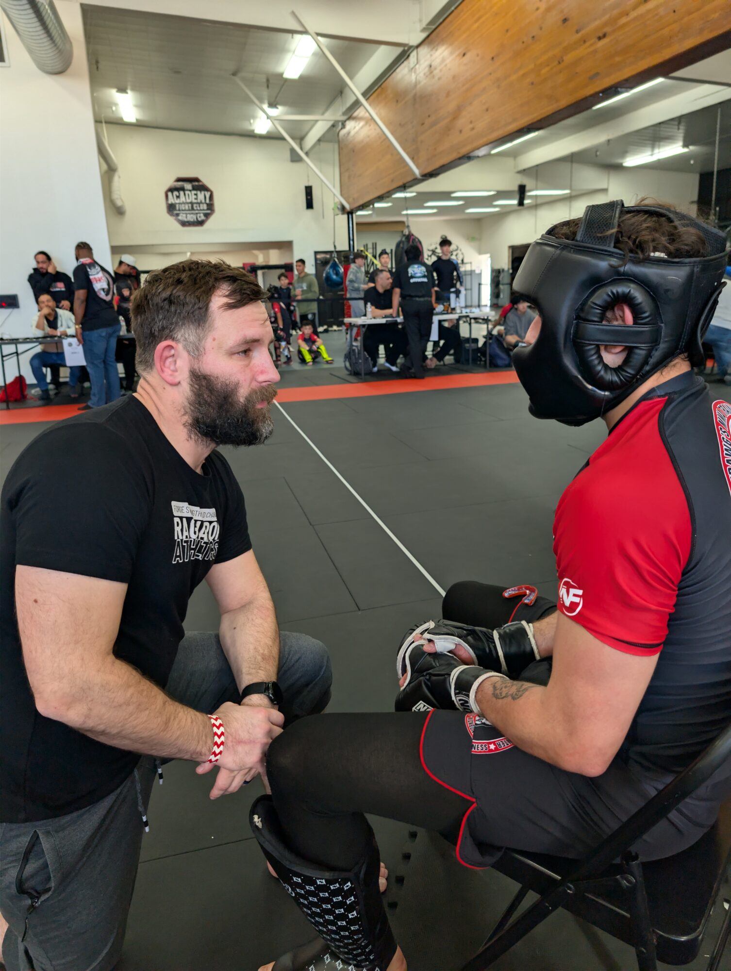 A man with a beard kneels facing a person in a black helmet and red shirt, inside a gym with people in the background.