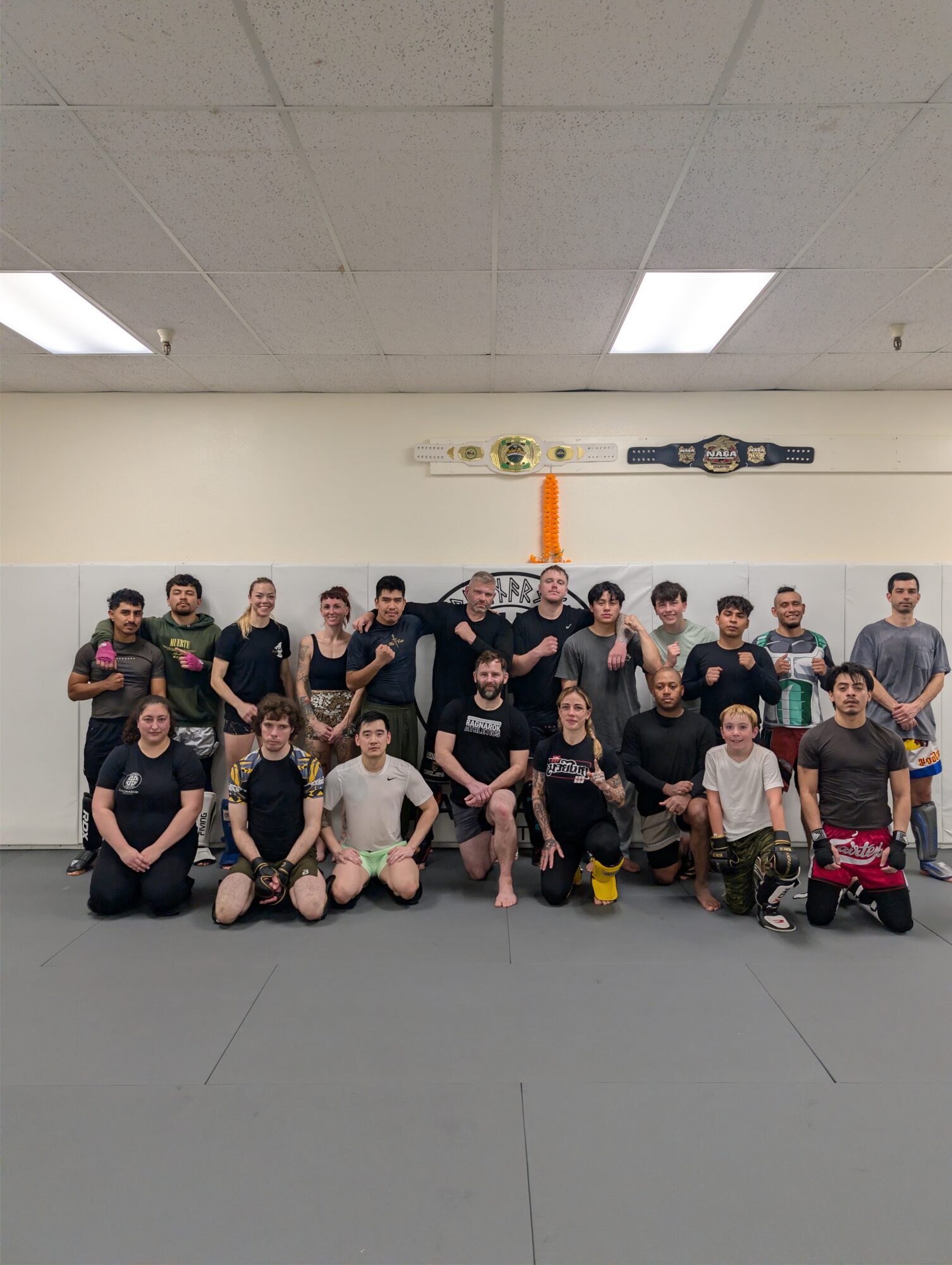 Group of people in a martial arts dojo, some holding belts, with a large belt display on the wall behind them.