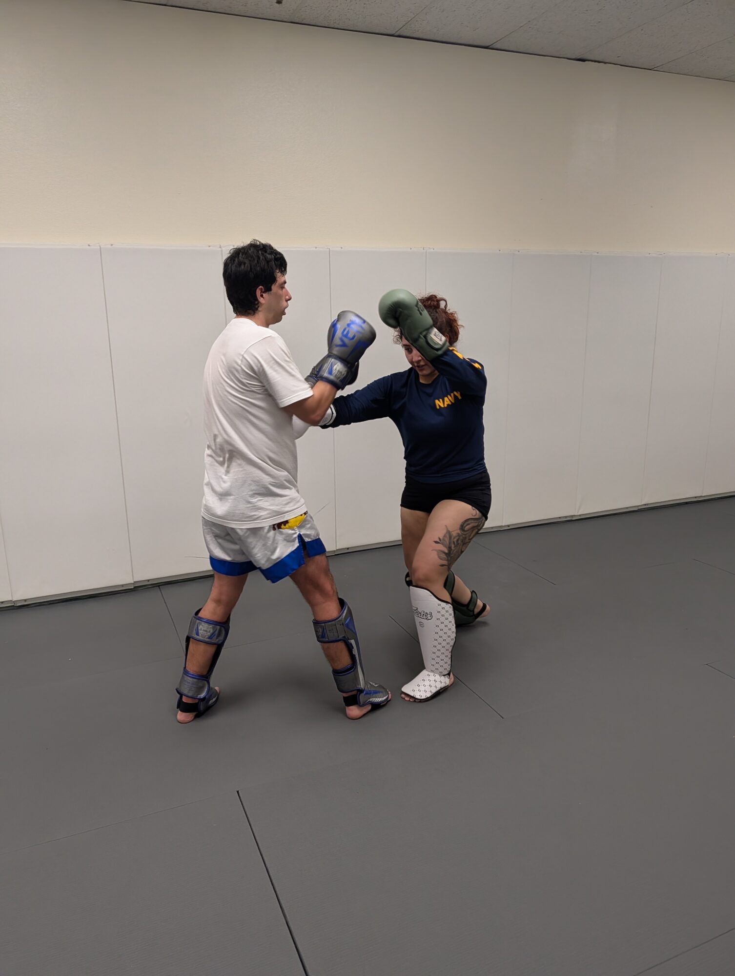Two people practicing boxing in a gym, wearing gloves and protective gear, facing each other in a fighting stance.