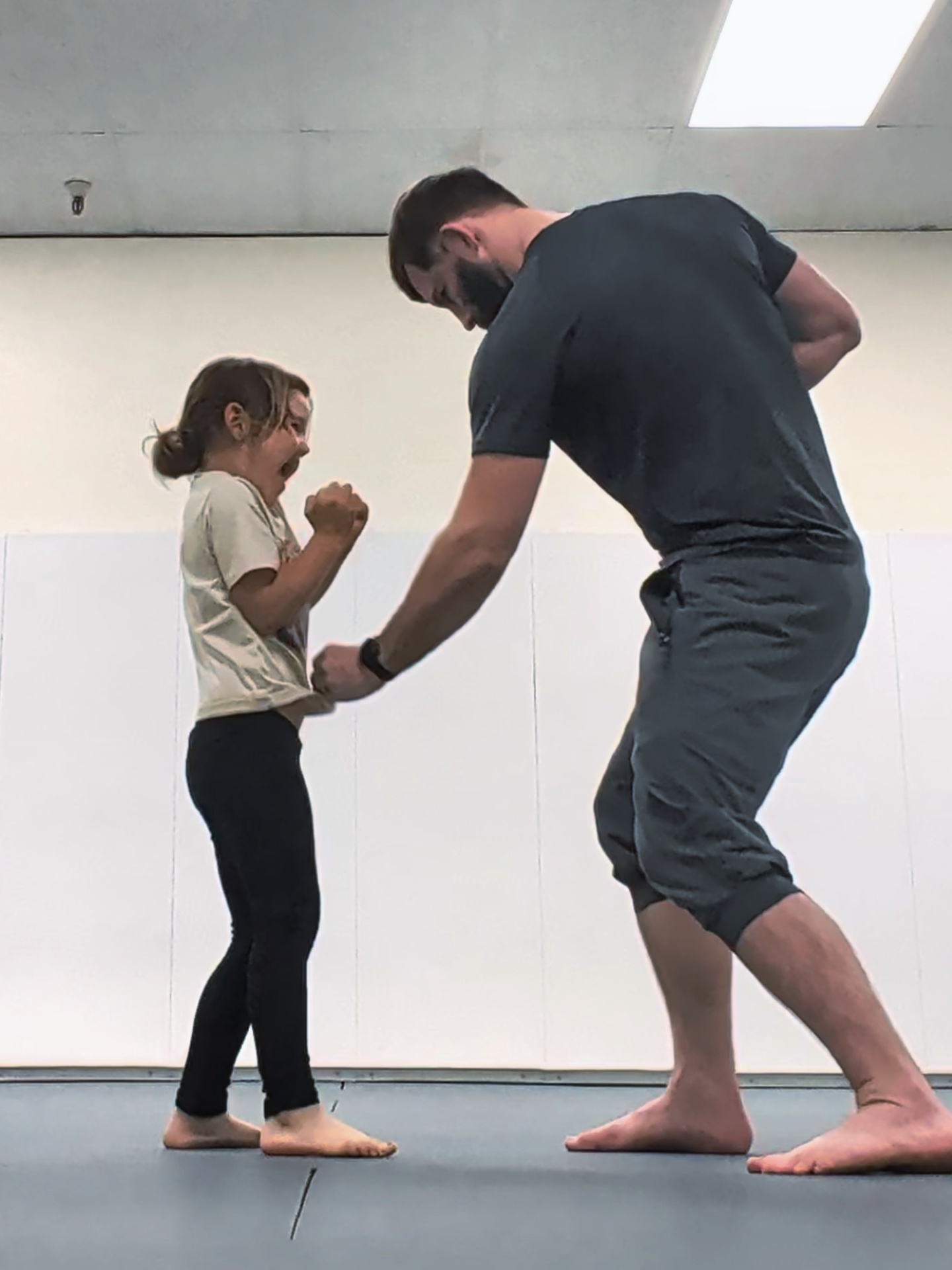 A man and a girl hold hands and face each other in a room with a white wall and ceiling light.