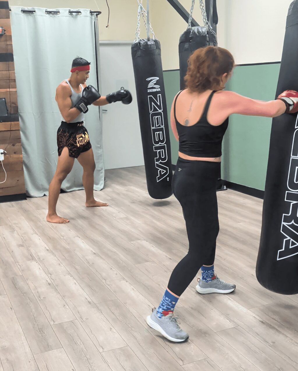 Two people training with punching bags in a gym, one man in shorts and gloves, one woman in workout clothes, both focused.