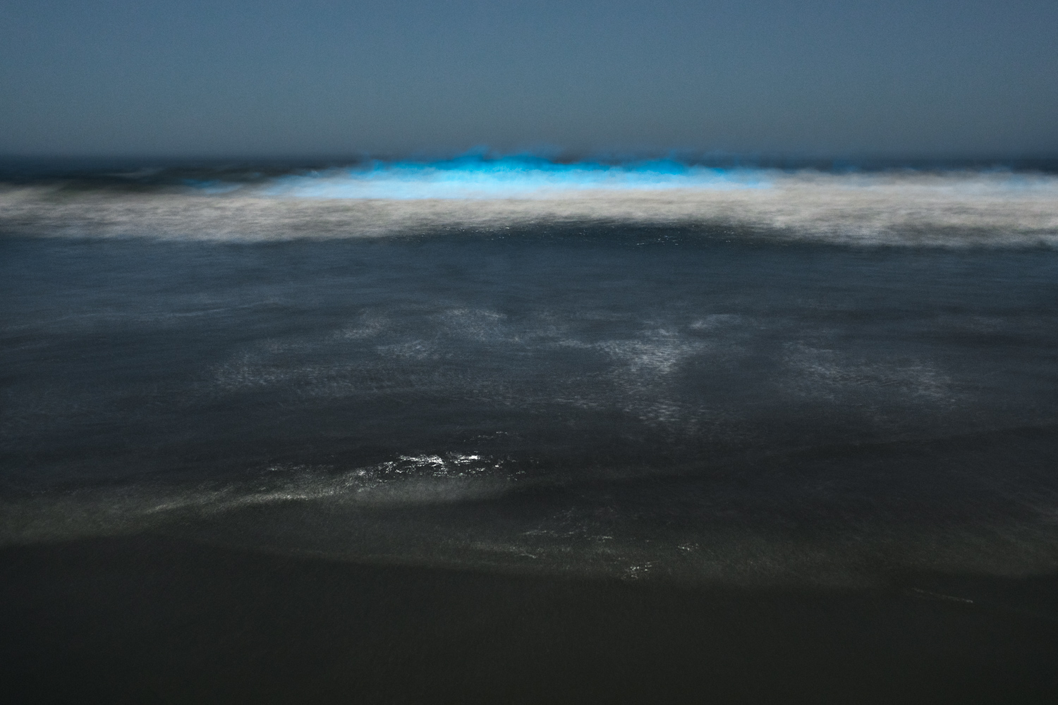 Dark ocean waves with a blue bioluminescent glow near the horizon, under a cloudy sky.