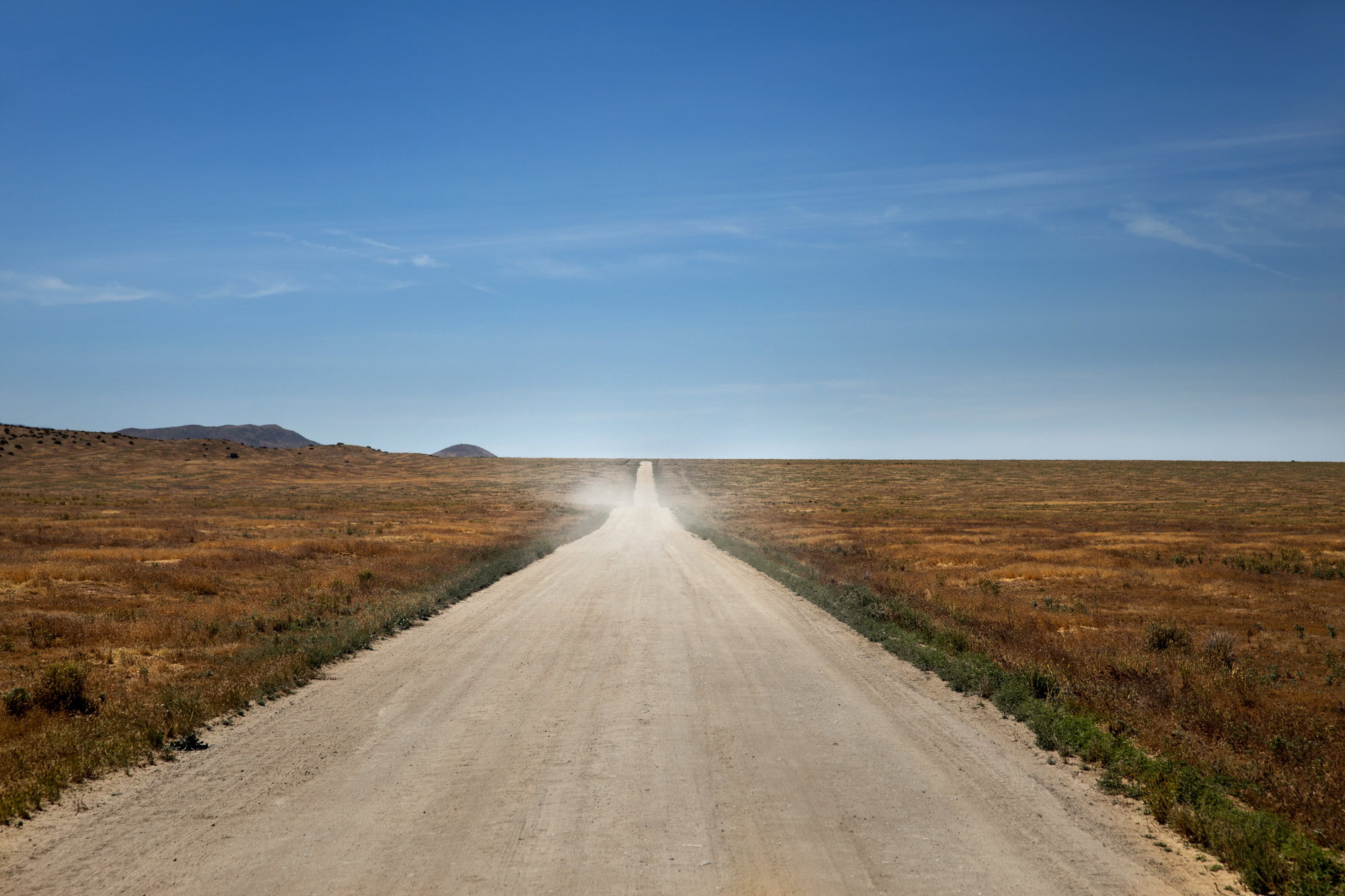 Straight dirt road extending into the horizon through open landscape with blue sky and distant hills.