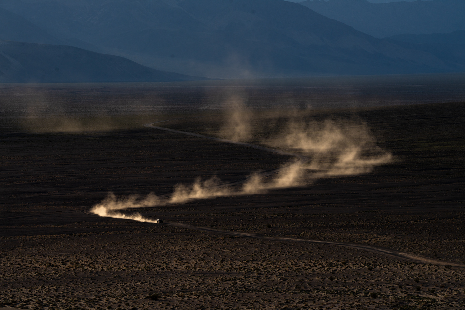 Dust clouds rise from dark, barren land with mountains in the background, under a cloudy sky.