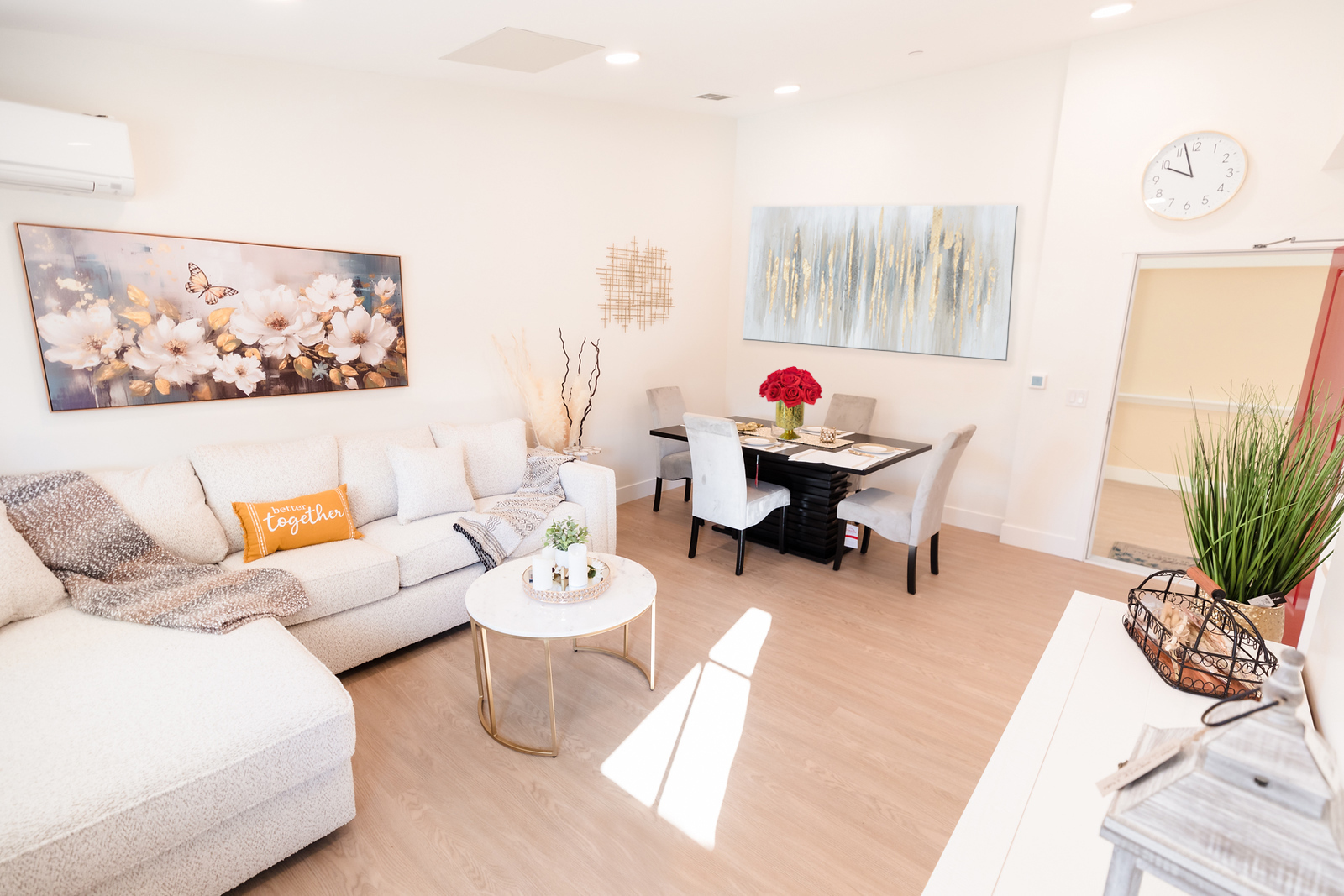 Living room with white sofa, coffee table, dining table with chairs, artwork, clock, and plants, bright and modern decor.