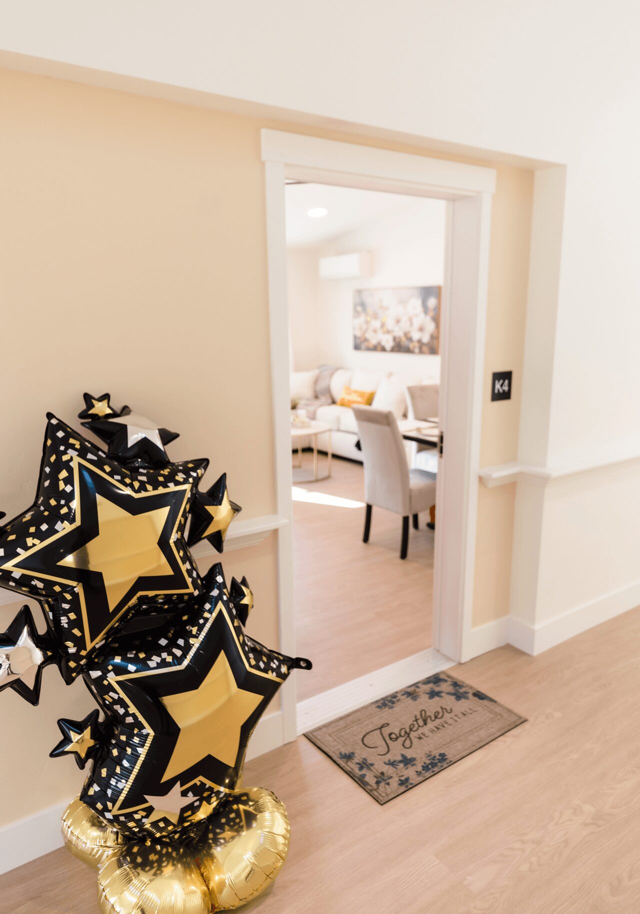 View into a living room through an open doorway, with a star-shaped balloon bouquet on the left and a welcome mat on the floor.