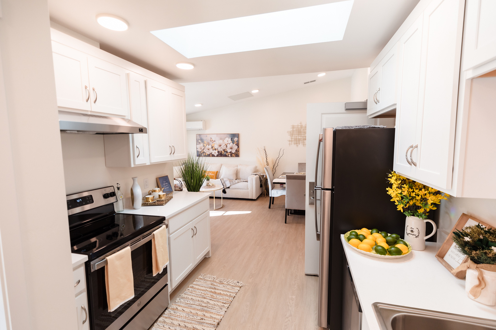 Kitchen with white cabinets, black refrigerator, stove, and a view into living room with sofa and artwork, ceiling skylight, and wooden floor.