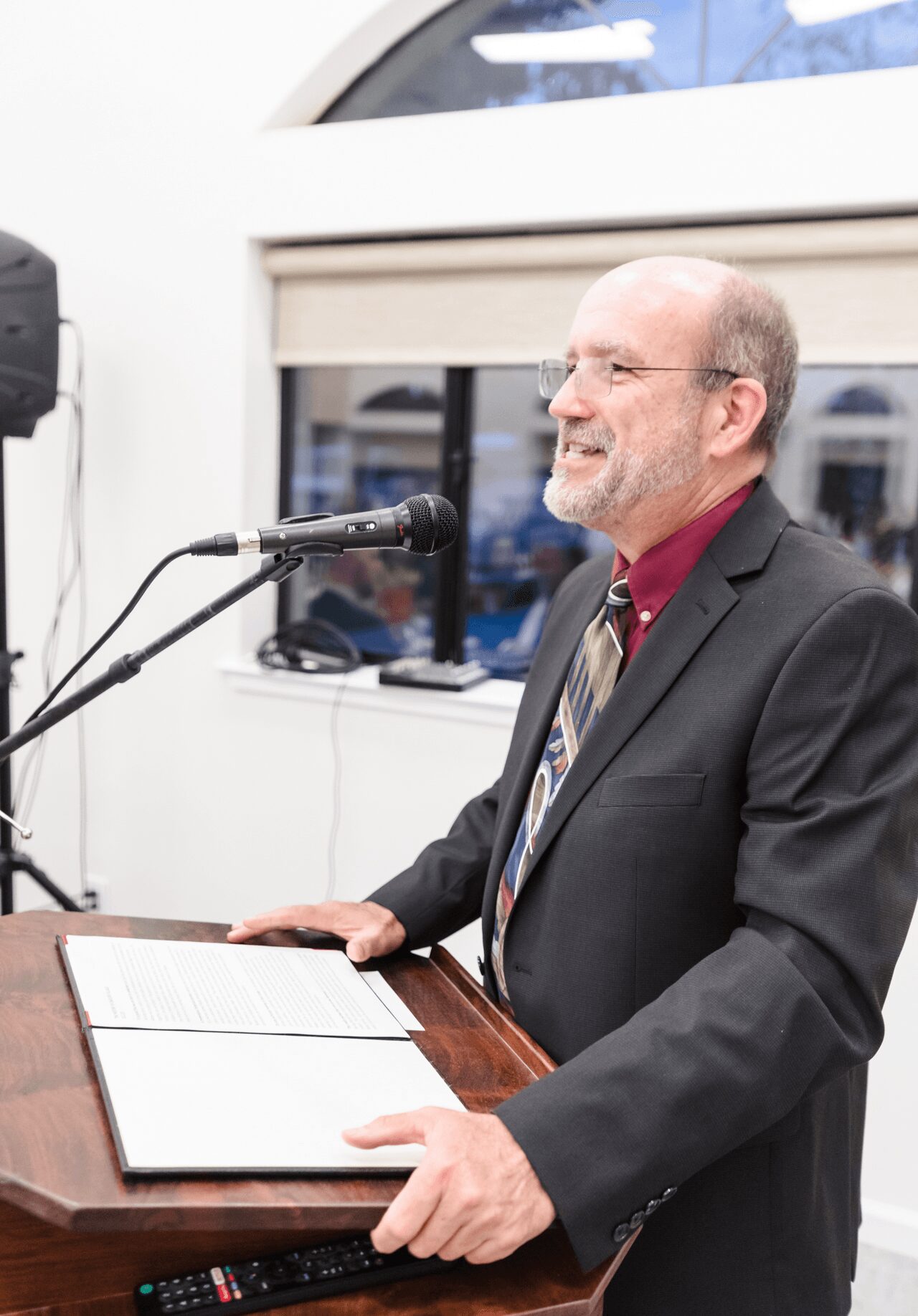 Man in suit speaking at a podium with a microphone, holding a paper, smiling, in a bright room with large windows.