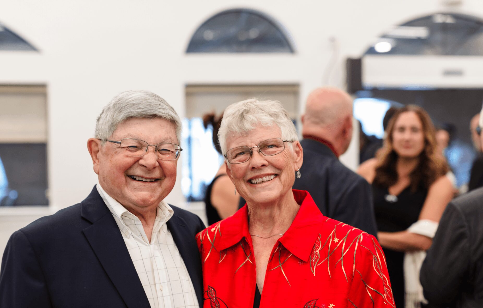 Two elderly people smiling and standing close together at an indoor event, with other people in the background.