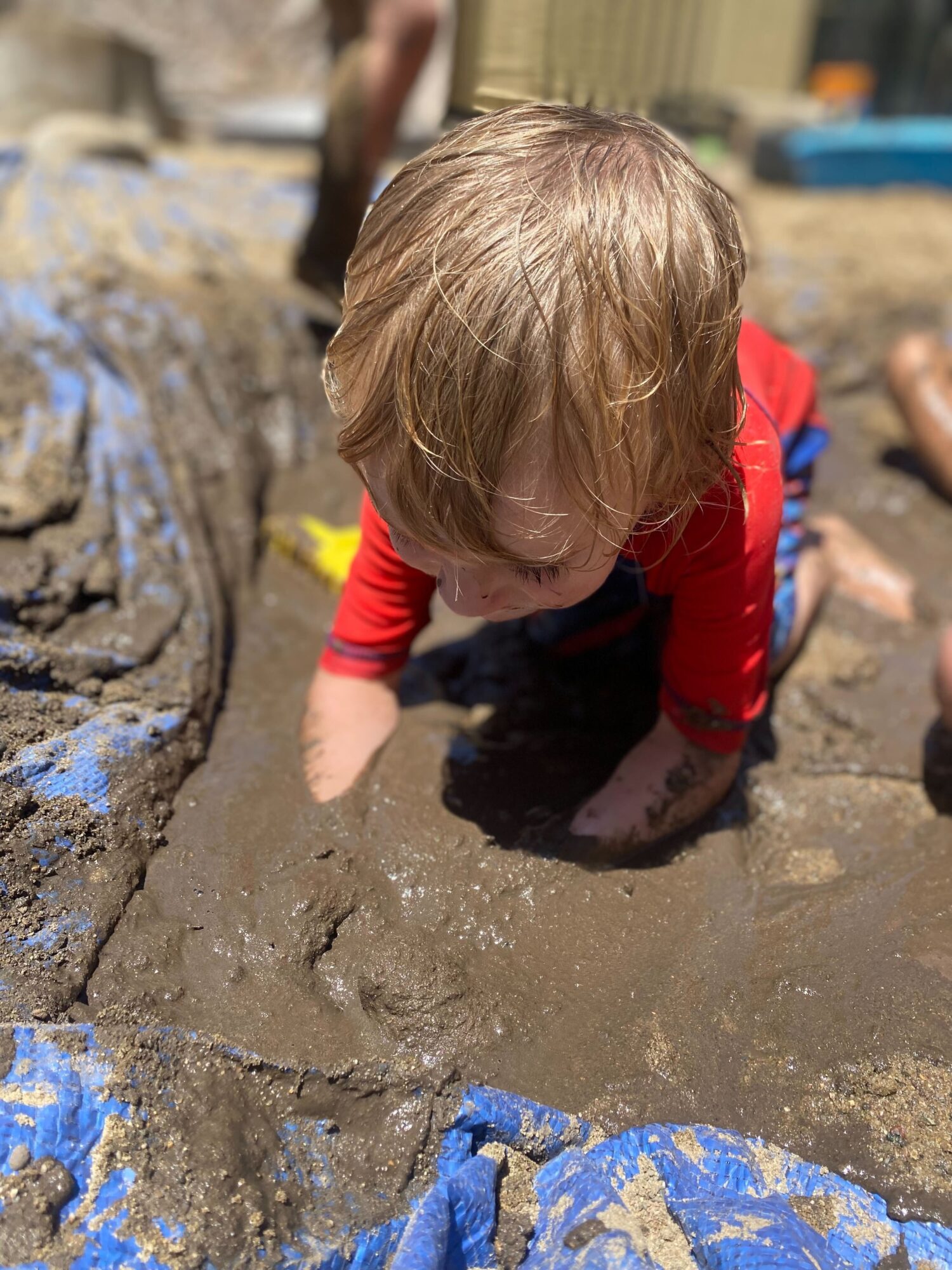Child with light brown hair in a red shirt crouching on muddy ground outdoors, playing with mud, with blurred background.