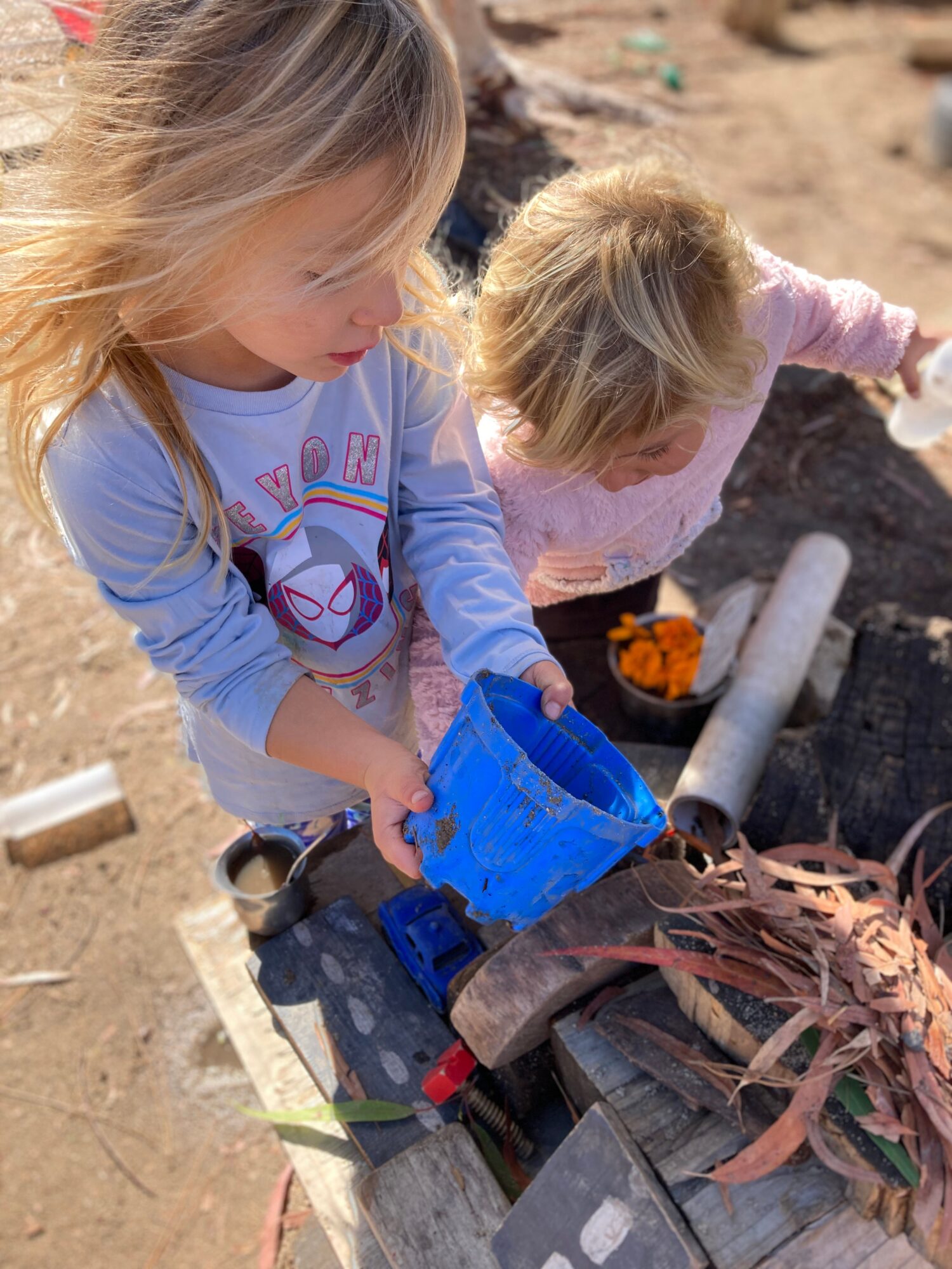 Two young children with blonde hair playing outdoors with a blue toy truck and natural objects on a wooden surface.