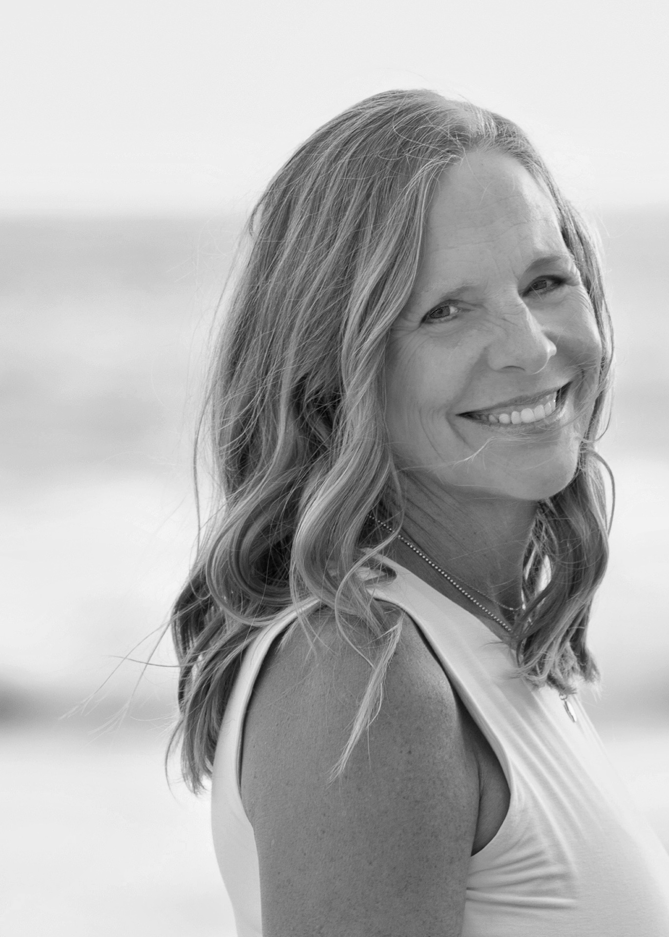 Smiling woman with wavy hair outdoors, background appears to be a beach or water, in black and white.
